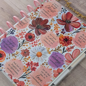 May include: A disc-bound planner featuring a floral pattern with red, orange, and purple flowers on a white background. The cover includes circular text boxes with Bible verses such as Philippians 4:13, Jeremiah 29:11, and Psalm 46:10. It has pink plastic discs.