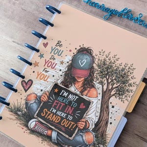 May include: A disc-bound notebook with a peach-colored cover featuring an illustration of a person wearing a baseball cap and holding a sign that reads, I'M NOT HERE TO FIT IN. I'M HERE TO STAND OUT. The cover also includes the text, Be YOU. Do YOU. For YOU.