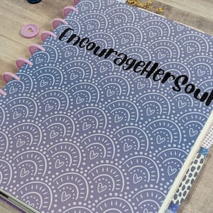 May include: A purple and white planner with a repeating pattern of white arches and hearts. The planner has a black vinyl sticker that says "Encourage Her Soul".