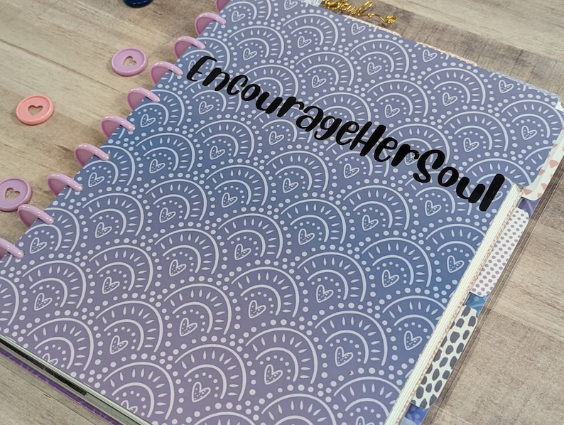 May include: A purple and white planner with a repeating pattern of white arches and hearts. The planner has a black vinyl sticker that says "Encourage Her Soul".