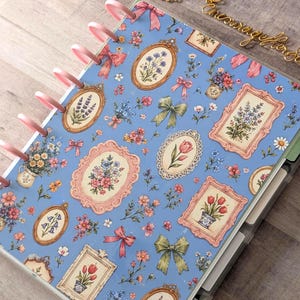 May include: A disc-bound planner with a light blue cover featuring a repeating pattern of floral bouquets in ornate frames, pink bows, and green ribbons. The planner is secured with pink plastic discs along the left side and includes internal dividers.