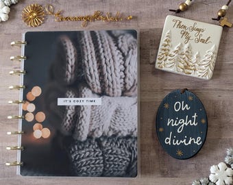 Op de afbeelding: Een witte planner met een gouden spiraalbinding en een foto van een knusse gebreide trui met de tekst "It's Cozy Time". Er zijn drie decoratieve ornamenten met de tekst "Then Sings My Soul", "Oh night divine", en "Encourage Your Soul".