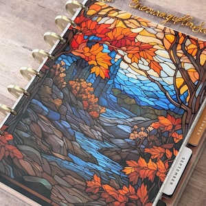 May include: A disc-bound planner featuring a stained-glass style cover illustration of a flowing stream surrounded by autumn trees with orange and red leaves. The planner includes gold-colored discs and visible monthly tab dividers for September, December, and November.