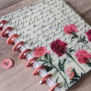 Op de afbeelding: Een notitieboek met schijfbinding en een omslag met vintage cursief handschrift en roze bloemenillustraties. Het notitieboek heeft metallic ros&eacute;gouden schijven langs de rug en bevat &eacute;&eacute;n losse, lichtroze hartvormige plastic schijf.
