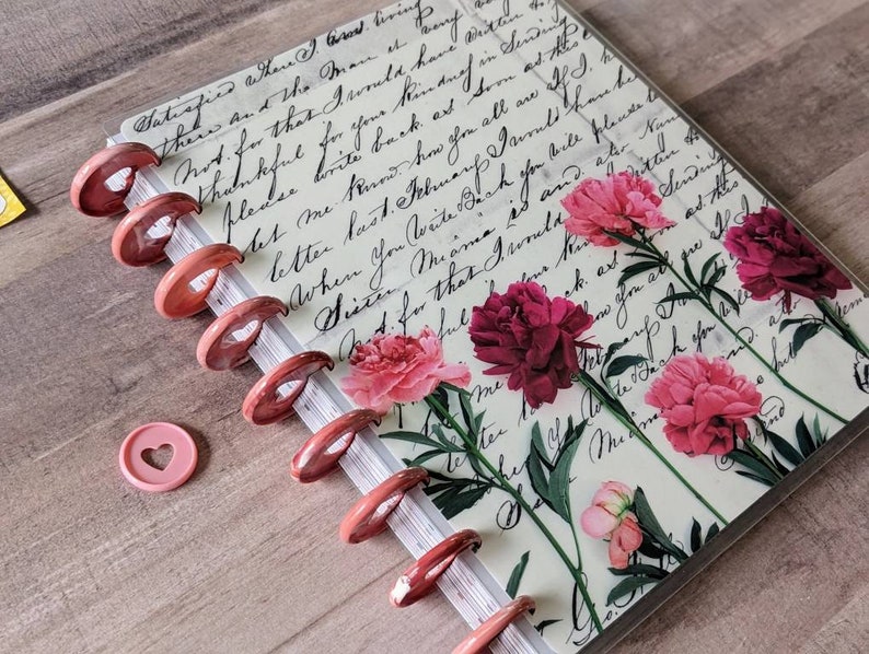 Op de afbeelding: Een notitieboek met schijfbinding en een omslag met vintage cursief handschrift en roze bloemenillustraties. Het notitieboek heeft metallic ros&eacute;gouden schijven langs de rug en bevat &eacute;&eacute;n losse, lichtroze hartvormige plastic schijf.