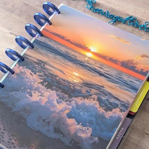 May include: A disc-bound planner featuring a cover image of an ocean sunset with crashing waves. The planner has dark blue plastic binding discs and includes monthly tab dividers labeled January, February, and March in white text.