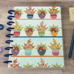 May include: A blue and white planner with a floral pattern. The planner has a clear cover and a spiral binding. The cover features a repeating pattern of colorful flower pots with flowers in them. The text "Enrich the Soul" is on a sticker in the upper right corner of the image.