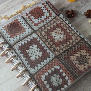 May include: A planner with a clear cover featuring a printed design of crocheted squares in shades of brown, white, and gray. The planner has gold-colored rings and a gold wire bookmark that says "Encourage Your Soul".