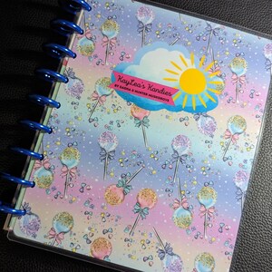 May include: A blue and purple planner with a clear plastic cover. The cover features a colorful pattern of cake pops with sprinkles and bows. The planner has a blue wire binding and the text "Kaylee's Kandies by Sasha & Hunter Musgrave" is printed on the cover.