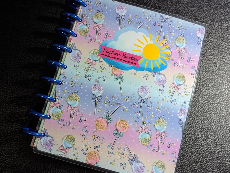 May include: A blue and purple planner with a clear plastic cover. The cover features a colorful pattern of cake pops with sprinkles and bows. The planner has a blue wire binding and the text "Kaylee's Kandies by Sasha & Hunter Musgrave" is printed on the cover.