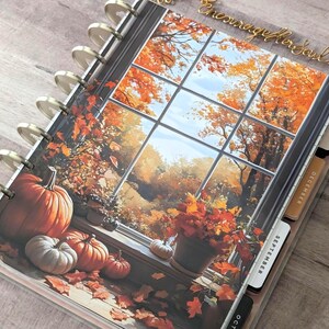 May include: A disc-bound planner featuring a cover illustration of a window view with autumn foliage, pumpkins, and a potted plant. The planner includes side tabs labeled September, October, and December, with gold-colored binding discs.