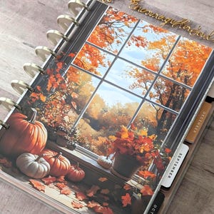 May include: A disc-bound planner featuring a cover illustration of a window looking out onto an autumn landscape with orange trees, pumpkins on a ledge, and fallen leaves. The planner includes tabbed dividers labeled September and December.