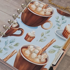 May include: A disc-bound planner with a light blue cover featuring a pattern of brown mugs filled with hot cocoa and marshmallows, cupcakes, and green leaves. The planner includes labeled monthly tabs for September, October, November, and December.