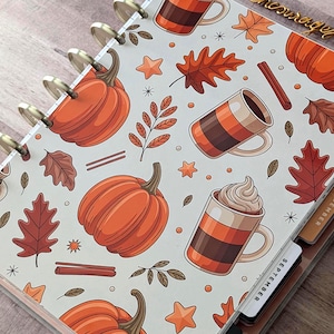 May include: A disc-bound planner featuring a light gray cover with an autumn-themed pattern of orange pumpkins, brown oak leaves, cinnamon sticks, star shapes, and mugs filled with coffee or whipped cream, with visible monthly tab dividers.