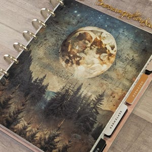 May include: A disc-bound planner featuring a cover with a full moon, a dark forest, and musical staff lines. The planner includes side tabs labeled September, December, and November, with the cursive text "Encourage her Soul" at the top right.