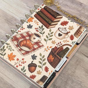 May include: A disc-bound planner featuring a cream cover with an autumn-themed pattern of books, a croissant, coffee mugs, a basket, acorns, and leaves. The planner includes monthly tab dividers labeled October, September, and December.