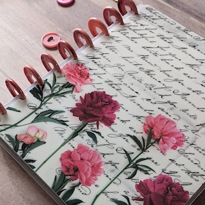 Op de afbeelding: Een notitieboek met schijfbinding en een omslag met een achtergrond van vintage cursief schrift en een bloemenpatroon van roze en donkerrode pioenen. Het notitieboek wordt bij elkaar gehouden door metallic koperkleurige plastic schijven.