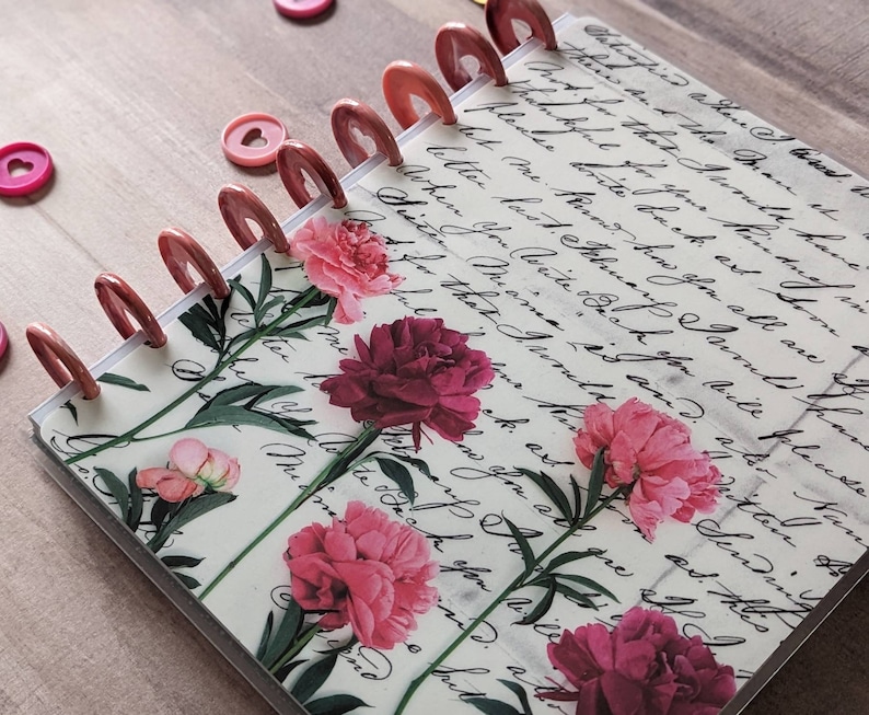 Op de afbeelding: Een notitieboek met schijfbinding en een omslag met een achtergrond van vintage cursief schrift en een bloemenpatroon van roze en donkerrode pioenen. Het notitieboek wordt bij elkaar gehouden door metallic koperkleurige plastic schijven.