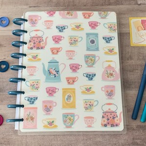 May include: A white planner with a clear plastic cover featuring a repeating pattern of teacups and teapots in various colors and designs. The planner has a ring binding system and is decorated with colorful heart-shaped paper clips.