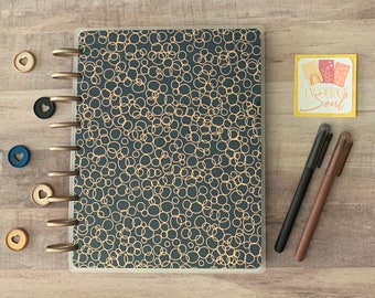 May include: A planner with a dark gray cover featuring a pattern of gold circles. The planner is open, revealing the binding rings and several colored discs. Two pens and a small card with the text "Encourage the Soul" are also present.