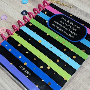 May include: A black and white striped planner with colorful stripes and gold polka dots. The planner has a pink binding and the text "Good, Better, Best... Never Let it Rest Until the Good is Better and the Better is Best!" in a blue oval. The planner is labeled "2023-2024".