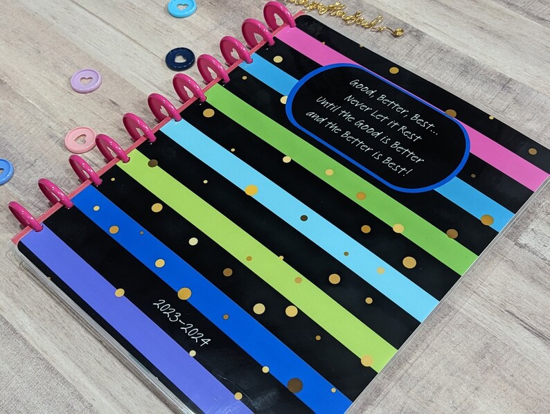 May include: A black and white striped planner with colorful stripes and gold polka dots. The planner has a pink binding and the text "Good, Better, Best... Never Let it Rest Until the Good is Better and the Better is Best!" in a blue oval. The planner is labeled "2023-2024".