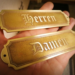 Könnte beinhalten: Zwei goldfarbene Metallplaketten mit verzierten Rändern. Die obere Plakette trägt die Aufschrift "Herren" und die untere Plakette die Aufschrift "Damen".