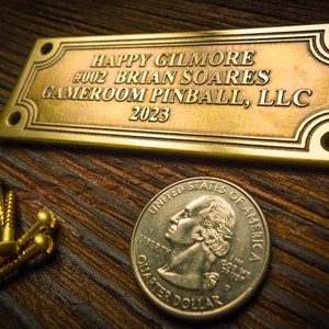 Puede incluir: Una placa de latón con el texto "HAPPY GILMORE #002 BRIAN SOARES GAMEROOM PINBALL, LLC 2023" sobre una superficie de madera. Hay tres tornillos dorados y una moneda de 25 centavos de dólar estadounidense junto a la placa.