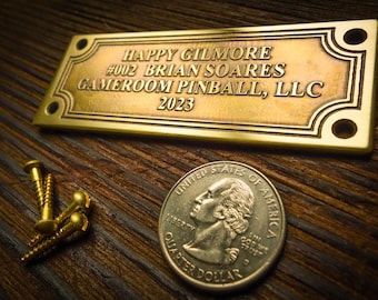 May include: A brass plaque with the text "HAPPY GILMORE #002 BRIAN SOARES GAMEROOM PINBALL, LLC 2023" on a wooden surface. There are three gold screws and a US quarter coin next to the plaque.