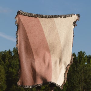 K&ouml;nnte beinhalten: Eine gewebte Decke mit Streifen in Rosa- und Beiget&ouml;nen, mit Fransen, vor blauem Himmel gehalten. Die Decke wird im Freien auf einer Wiese mit B&auml;umen im Hintergrund pr&auml;sentiert.