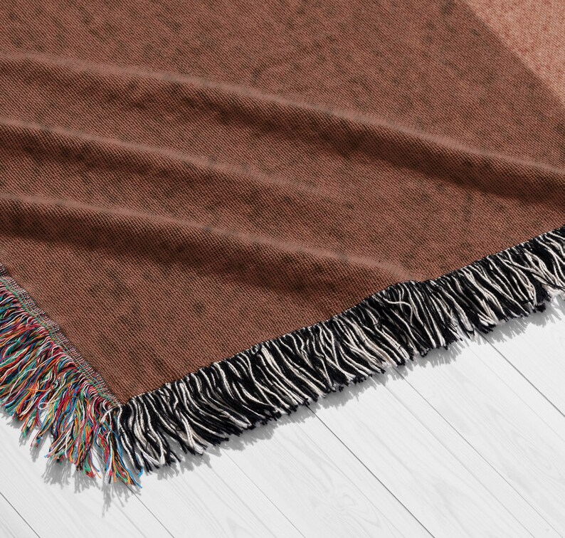 K&ouml;nnte beinhalten: Eine gewebte braune Wurkdecke mit einem gesprenkelten Muster. Die Decke hat einen Fransenrand mit schwarzen und wei&szlig;en Quasten und einen bunten Fransen an einer Ecke. Die Decke wird auf einer wei&szlig;en Holzoberfl&auml;che pr&auml;sentiert.