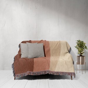 K&ouml;nnte beinhalten: Eine gewebte Wurfadecke in Rost-, Pfirsich- und Beiget&ouml;nen bedeckt ein kleines Sofa. Zwei graue quadratische Kissen liegen auf dem Sofa. Die Decke hat einen Fransenrand. Eine Topfpflanze in einem goldenen St&auml;nder befindet sich rechts.