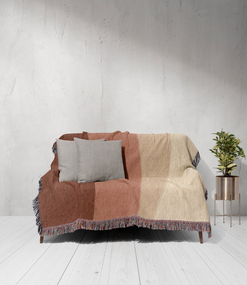 K&ouml;nnte beinhalten: Eine gewebte Wurfadecke in Rost-, Pfirsich- und Beiget&ouml;nen bedeckt ein kleines Sofa. Zwei graue quadratische Kissen liegen auf dem Sofa. Die Decke hat einen Fransenrand. Eine Topfpflanze in einem goldenen St&auml;nder befindet sich rechts.
