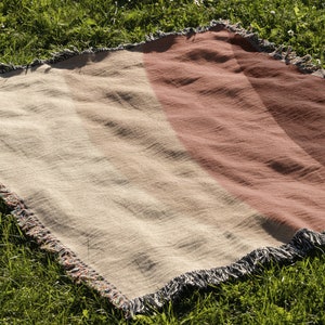 K&ouml;nnte beinhalten: Eine gewebte Decke mit Streifen in Beige- und Rotbraunt&ouml;nen und einem Fransenrand. Die Decke liegt auf gr&uuml;nem Gras, was auf eine Verwendung im Freien hindeutet. Das Design zeigt Streifen.
