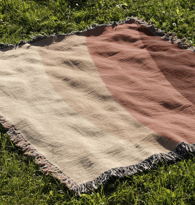 K&ouml;nnte beinhalten: Eine gewebte Decke mit Streifen in Beige- und Rotbraunt&ouml;nen und einem Fransenrand. Die Decke liegt auf gr&uuml;nem Gras, was auf eine Verwendung im Freien hindeutet. Das Design zeigt Streifen.