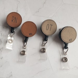 May include: Four brown and beige leather retractable badge holders with black clips and clear straps. The holders have the letters "RN" and "OT" printed on them.