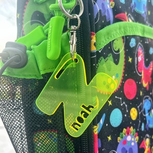 May include: A neon green acrylic keychain in the shape of the letter "N" with the name "noah" printed in yellow. The keychain is attached to a backpack with a dinosaur and space-themed pattern.