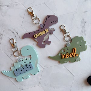 Może przedstawiać: Trzy breloki w kształcie dinozaur&oacute;w z napisami "Landon", "Noah" i "Noah". Dinozaury są fioletowe, zielone i niebieskie.
