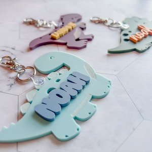 Może przedstawiać: Trzy niebieskie, zielone i fioletowe breloczki w kształcie dinozaur&oacute;w z imionami Noah, Noah i Lily. Breloczki są wykonane z plastiku i mają uroczy, kresk&oacute;wkowy design.