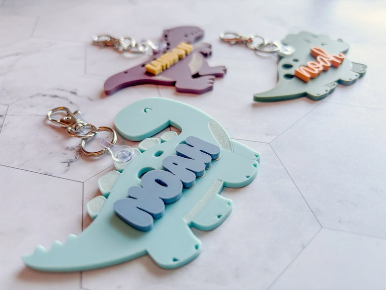 Może przedstawiać: Trzy niebieskie, zielone i fioletowe breloczki w kształcie dinozaur&oacute;w z imionami Noah, Noah i Lily. Breloczki są wykonane z plastiku i mają uroczy, kresk&oacute;wkowy design.