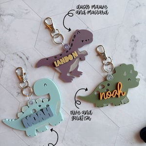 Może przedstawiać: Trzy breloki w kształcie dinozaur&oacute;w z r&oacute;żnymi nazwami i kolorami. Pierwszy brelok jest fioletowy z imieniem Landon, drugi jest jasnoniebieski z imieniem Noah, a trzeci jest zielony z imieniem Noah.