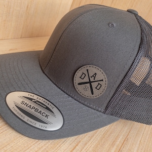 May include: A gray and black baseball cap with a mesh back. The front of the cap has a black leather patch with a baseball bat and the word "DAD" in a circle.