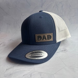 May include: A navy blue and white trucker hat with a brown leather patch that says "DAD" on it. The hat has a mesh back and a snap closure. The hat is made by Yupoong and is a snapback style.