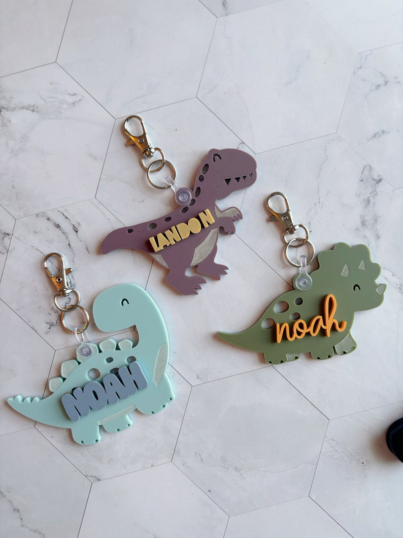 Może przedstawiać: Trzy breloki w kształcie dinozaur&oacute;w z napisami "Landon", "Noah" i "Noah". Dinozaury są fioletowe, zielone i niebieskie.