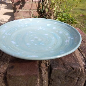 Peut inclure: Une assiette creuse ronde en céramique, émaillée bleu clair avec des mouchetures blanches. L'assiette présente un motif en spirale et est adaptée pour servir des aliments. L'assiette est posée sur une surface en briques.
