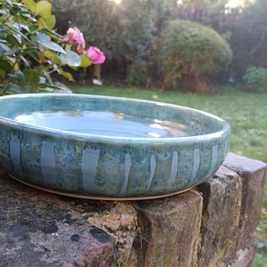 May include: A handmade, shallow ceramic bowl with a teal and blue glaze. Vertical, lighter blue stripes decorate the exterior. The bowl is set on a brick surface, with a blurred garden background.