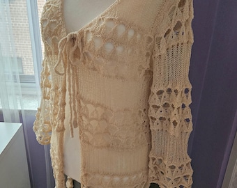 May include: A beige crochet cardigan with a tie at the neckline and a loose, flowy fit. The cardigan has a delicate, open-weave pattern.