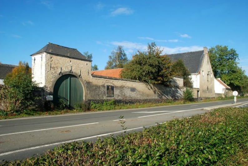 May include: A stone building with a green wooden door and a stone wall in front of it. The building has a gray roof and a white facade. There is a road in front of the building and a hedge on the right side of the road.