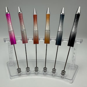 May include: Six pens with silver accents and different colored bodies. The pens are arranged in a row on a clear plastic stand. The colors of the pens are pink, white, orange, gold, gray, and black.