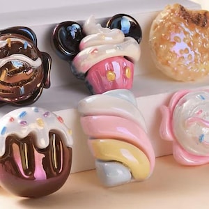 May include: A collection of colorful, iridescent ceramic food-themed decorative objects. The items include a cupcake with Mickey Mouse ears, a donut, a marshmallow, and other sweet treats. The objects are arranged on a white surface.